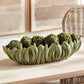 The Winslow Bowl Collection by Napa Home & Garden, featuring a decorative green leaf pattern and reactive-glazed stoneware, displays artificial moss balls as a stunning centerpiece on a light wooden table with abstract art in the background.