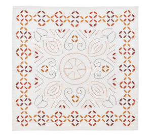 The Malta Napkin Set by Kim Seybert features square white napkins with symmetrical, hand-stitched geometric and floral patterns in orange, brown, and yellow, celebrating traditional cutwork techniques.