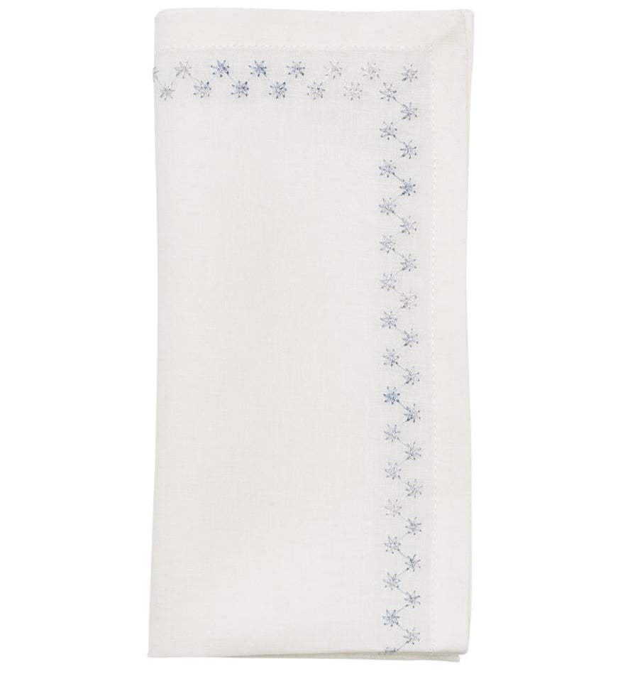 The Starry Night Napkin Set by Kim Seybert features folded white linen napkins with shimmering metallic blue floral embroidery along the border, adding a delicate, elegant touch to any table setting.