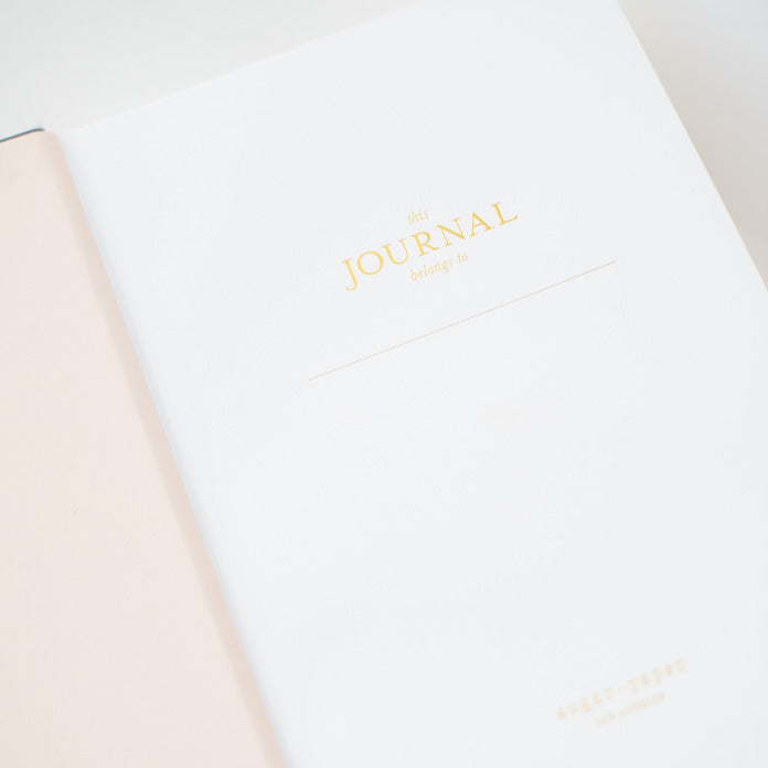 Open the Pink Gingham Tailored Journal by Sugar Paper to reveal gold text on the first page that reads “this JOURNAL belongs to,” with a blank space underneath for your name. Featuring a light pink book cloth hardcover on the left and lined pages inside, this is the perfect journal for personal reflections.
