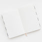An open Sugar Paper Essential Flax Dot Journal with blank white pages and a beige ribbon bookmark lies on a white surface.
