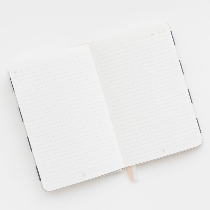 The Sugar Paper Essential Chambray Stripe Journal, featuring lined pages and a ribbon bookmark, rests on a white surface.
