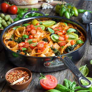 A Hestan NanoBond Titanium Sauté Pan, 5qt, filled with pasta, cherry tomatoes, green tomatoes, broccoli, and fresh basil sits on a wooden table surrounded by spices and fresh vegetables.