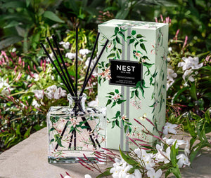 The Nest Indian Jasmine Decorative Reed Diffuser stands next to its floral box on a stone surface, surrounded by white jasmine blooms and filled with premium fragrance oils.