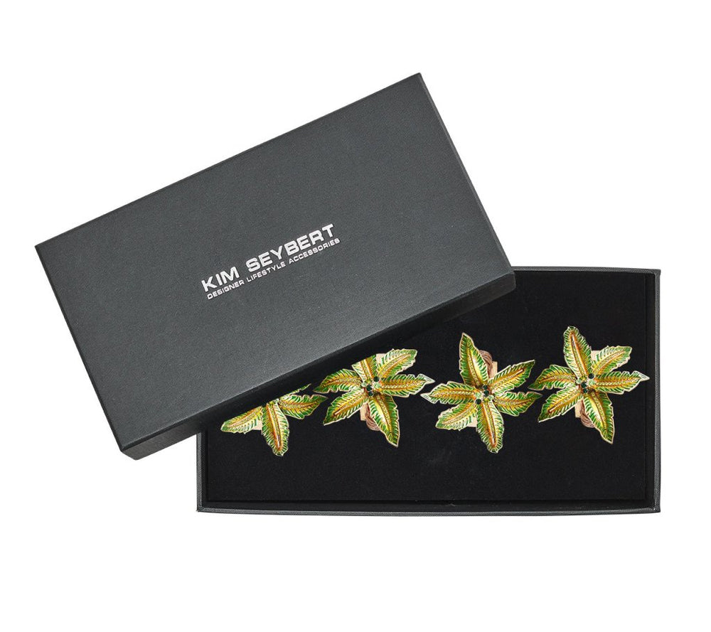 A black Kim Seybert box, lid ajar, displays a set of four Palm Coast Napkin Rings—nature-inspired table decor in green, gold, and brown.