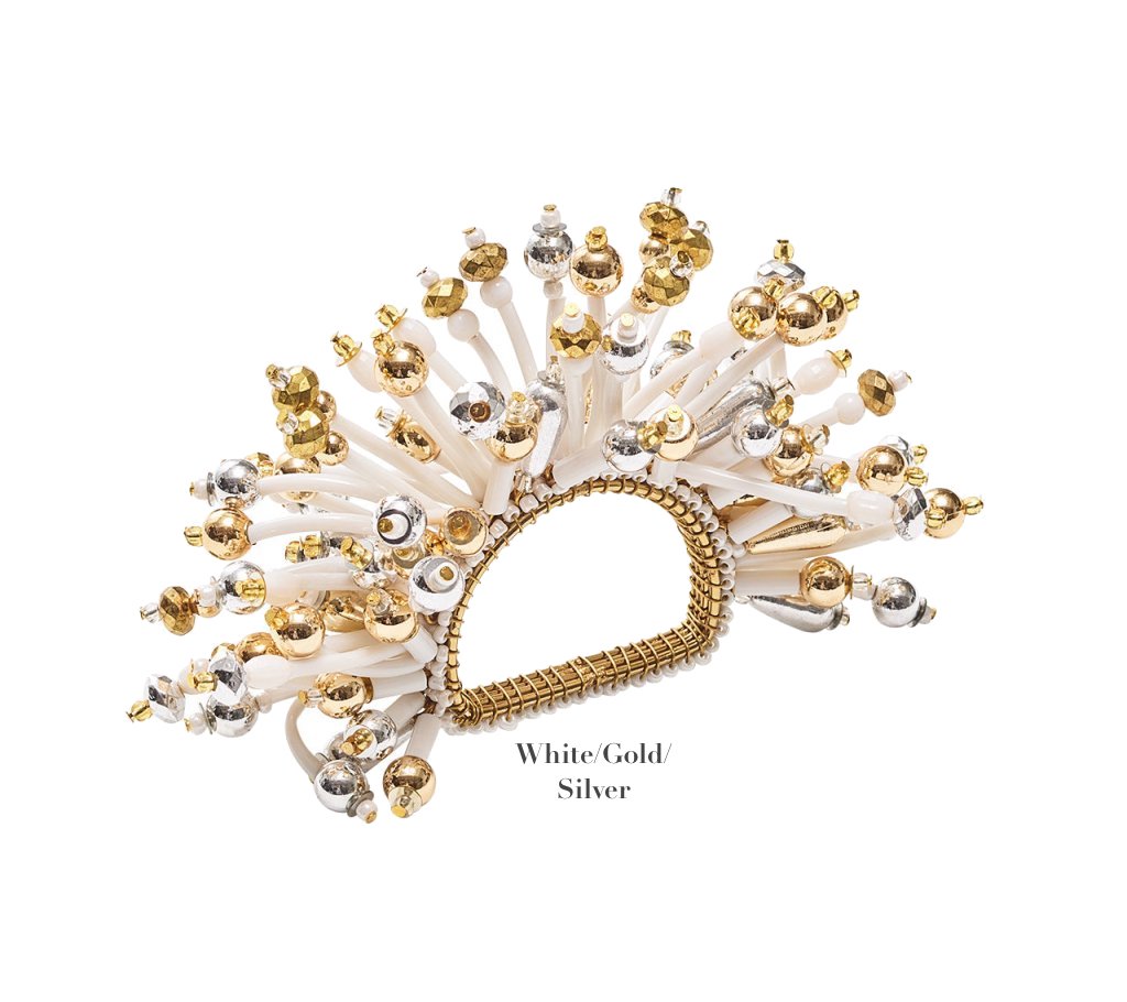 The Kim Seybert Fun Burst Napkin Ring features white, gold, and silver elements in a fan shape with metallic beads, silicone tubes, and a woven base. Text reads "White/Gold/Silver.