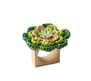 The Kim Seybert Pareo Napkin Ring features a decorative green and beige raffia-style flower made from woven material attached to a wooden ring.