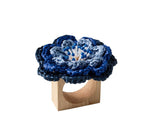 The Pareo Napkin Ring by Kim Seybert features a square wooden base topped with a blue hand-crocheted flower, bringing unique artisanal charm to your table setting.