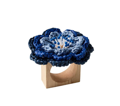 The Pareo Napkin Ring by Kim Seybert features a square wooden base topped with a blue hand-crocheted flower, bringing unique artisanal charm to your table setting.