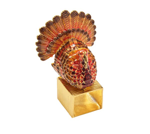 A Kim Seybert Turkey Trot Napkin Ring featuring a gold-toned square base topped with a metallic, multi-colored turkey figurine for Thanksgiving table decor.
