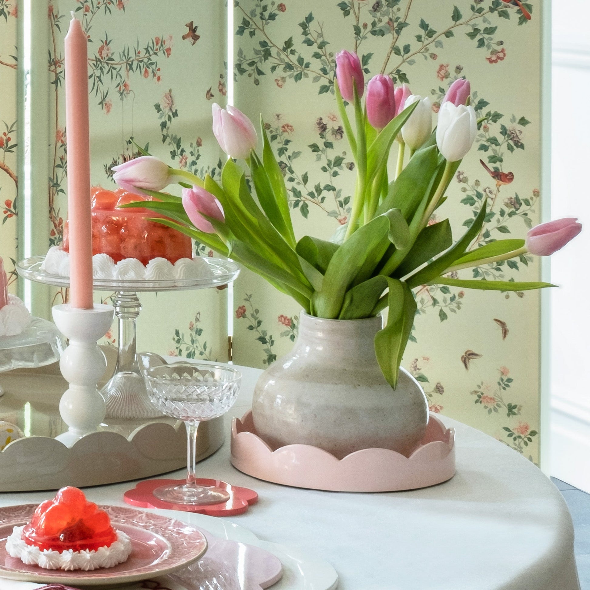 A vase with pink and white tulips sits on a round table set with pink candles, glassware, desserts, and Addison Ross's Round Scalloped Lacquer Tray Collection, all against a floral wallpaper background.