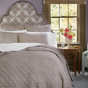 A neatly made bed with the John Robshaw Nandi Coffee Quilt in taupe and white pillows sits by a window; a vase of pink flowers and a glass of water rest on a wooden side table.