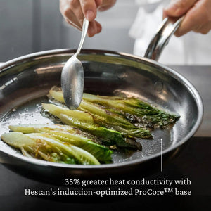 A person uses a spoon to cook leafy greens in a Hestan NanoBond Titanium Skillet, 12.5", with text highlighting its exceptional heat conductivity and metal utensil safe design.