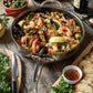 A pan of seafood paella with shrimp, mussels, and lemon wedges rests on a wooden table in the Hestan NanoBond Titanium Essential Pan, 5qt, surrounded by bread, olives, fresh herbs, and glasses of red wine.