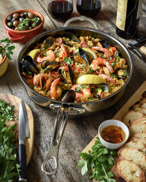 A pan of seafood paella with shrimp, mussels, and lemon wedges rests on a wooden table in the Hestan NanoBond Titanium Essential Pan, 5qt, surrounded by bread, olives, fresh herbs, and glasses of red wine.