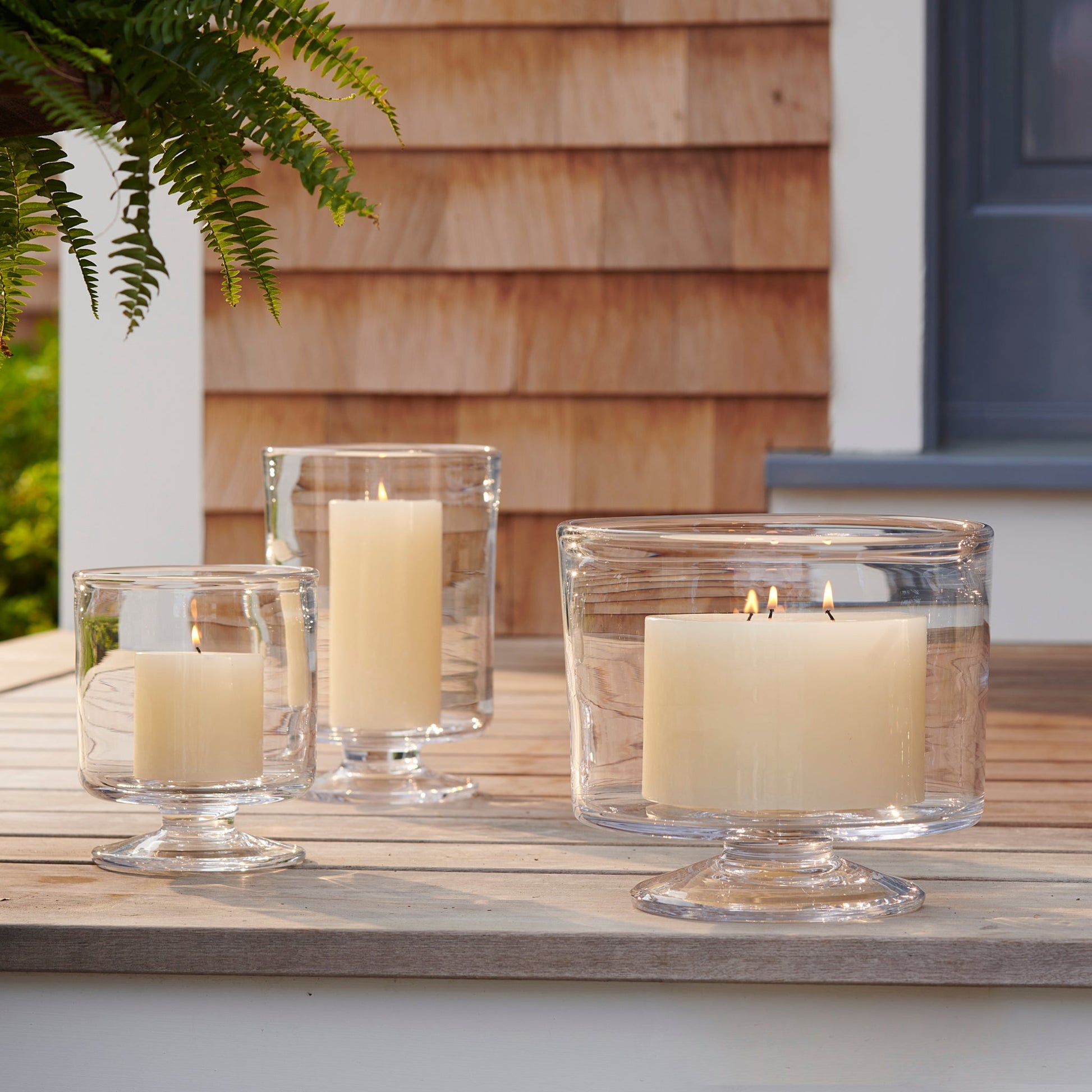 Three Nantucket Hurricane Collection candle holders by Simon Pearce, each with a white pillar candle, are displayed on an outdoor wooden table beside a shingled Nantucket-style house.