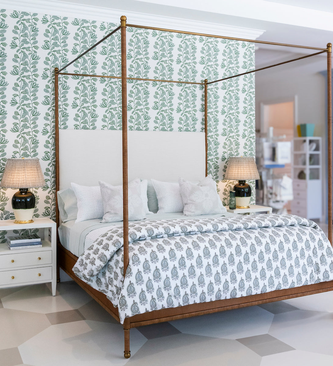 A bedroom featuring the Julian Chichester Rita Cane Wrap Four Poster Bed, white and patterned bedding, two bedside tables each with a lamp, and green leafy wallpaper behind the bed.