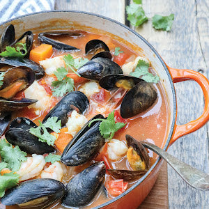 Coastal Kitchen by Simon & Schuster features a vibrant orange pot brimming with seafood stew—packed with mussels, shrimp, tomatoes, and vegetables, and topped with fresh cilantro on a wooden table—making it the ideal choice for anyone who loves coastal living and preparing wholesome seafood dishes at home.