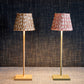 Two Zafferano x Fermoie Poldina Lampshade Collection table lamps by Zafferano America, featuring slender stands and hand-made lampshades, one brown and one pink, rest on a textured surface against a dark woven backdrop.