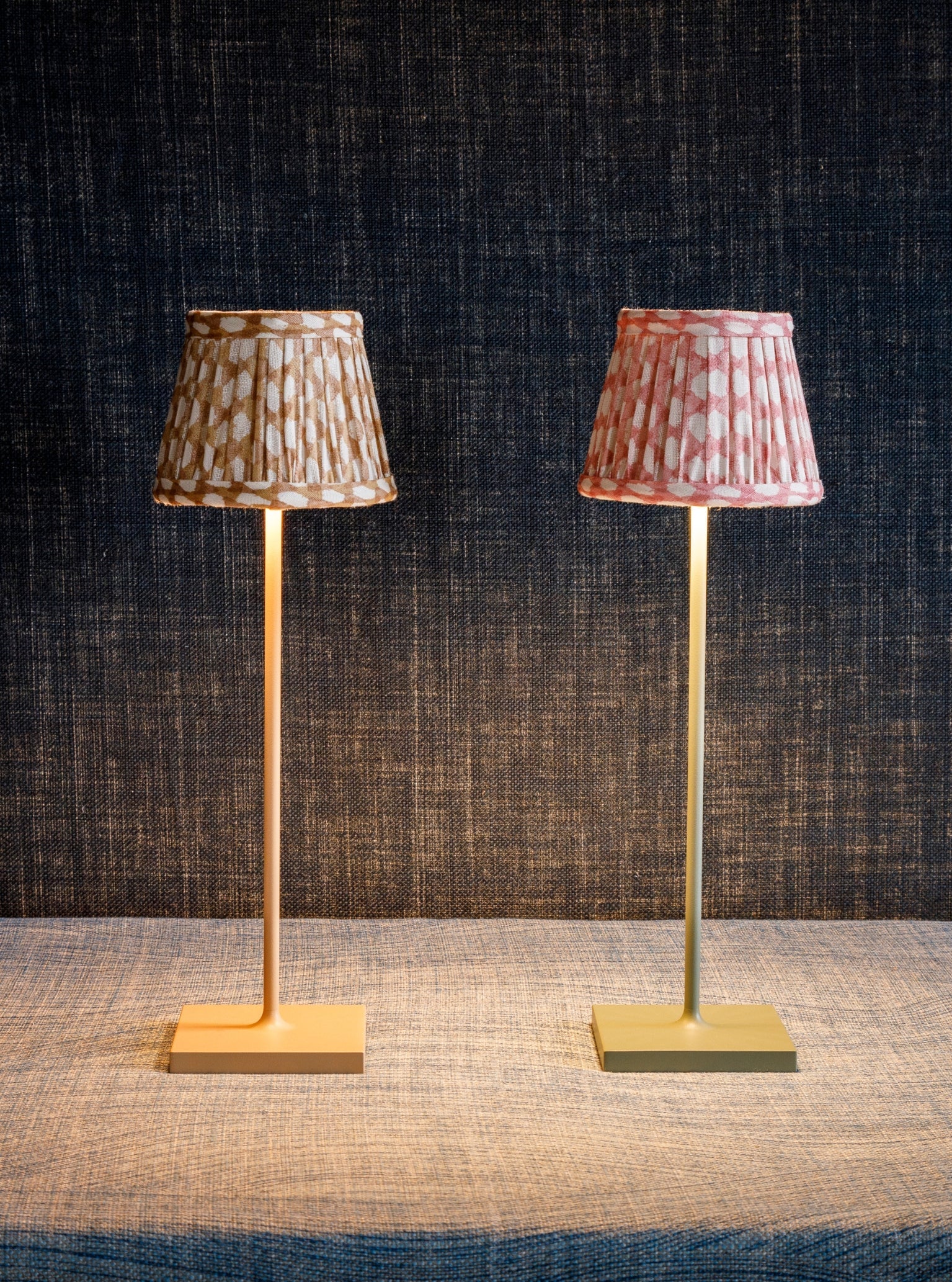 Two Zafferano x Fermoie Poldina Lampshade Collection table lamps by Zafferano America, featuring slender stands and hand-made lampshades, one brown and one pink, rest on a textured surface against a dark woven backdrop.