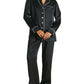 A person wearing Olivia von Halle’s Coco Silk Pajamas in black with white piping—a long-sleeve button-up top and matching pants—stands barefoot.