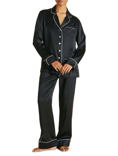 A person wearing Olivia von Halle’s Coco Silk Pajamas in black with white piping—a long-sleeve button-up top and matching pants—stands barefoot.