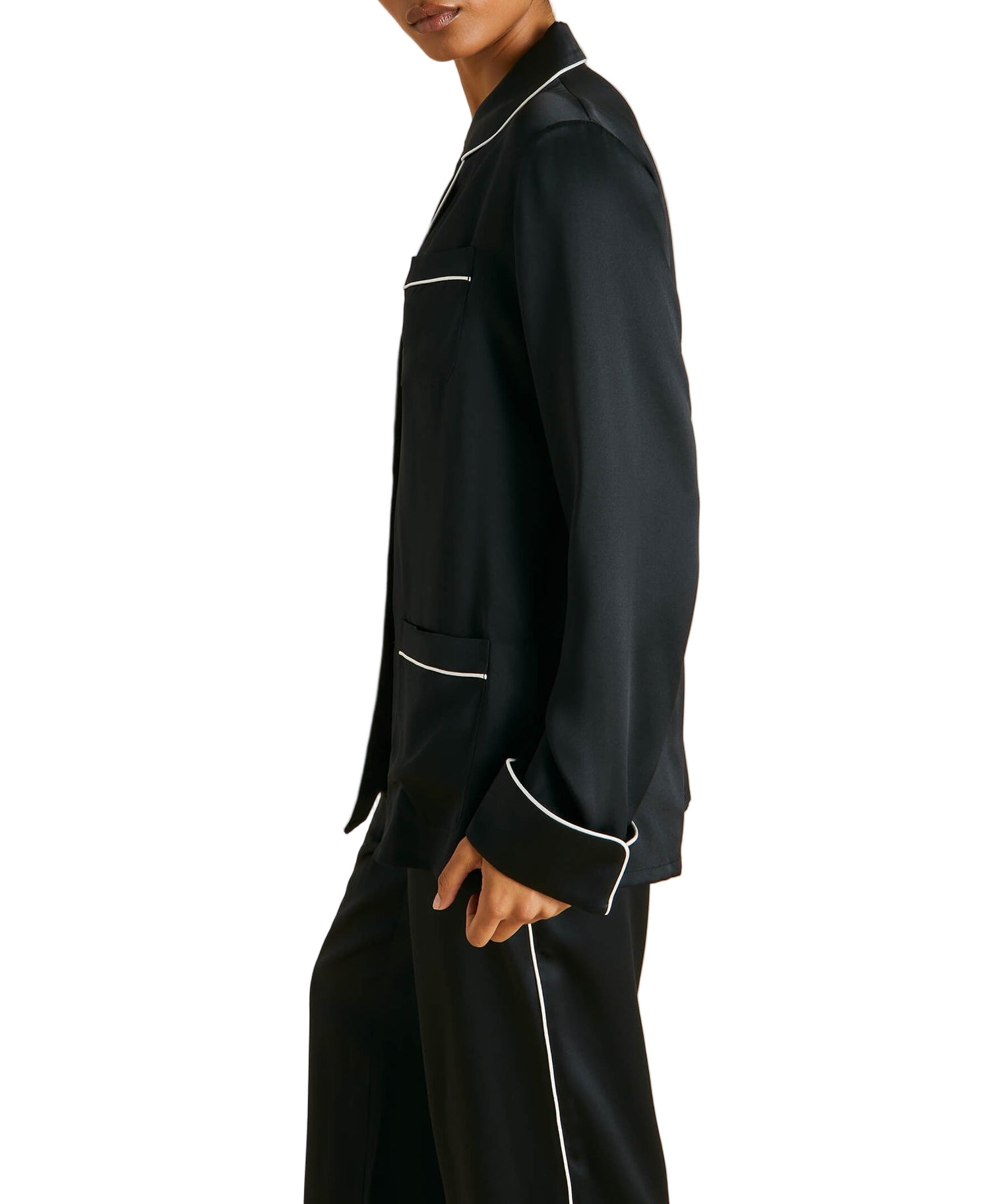 A model in Olivia von Halle’s Coco Silk Pajamas—black with white piping—appears from shoulder to mid-thigh, turned slightly sideways against a white background.