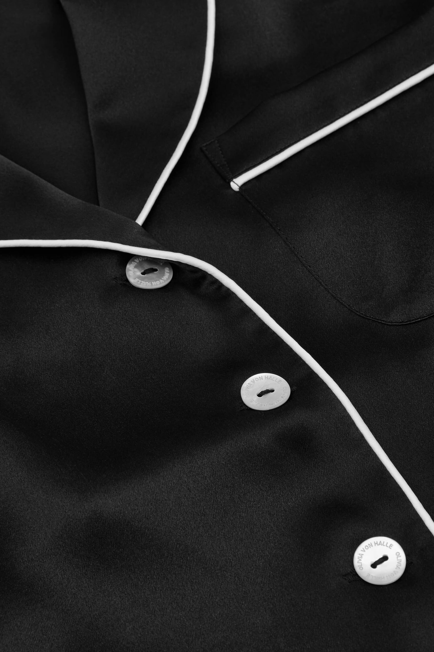 Close-up of an Olivia von Halle Coco Silk Pajamas black satin shirt with white piping, white buttons, and a chest pocket—luxurious eveningwear.