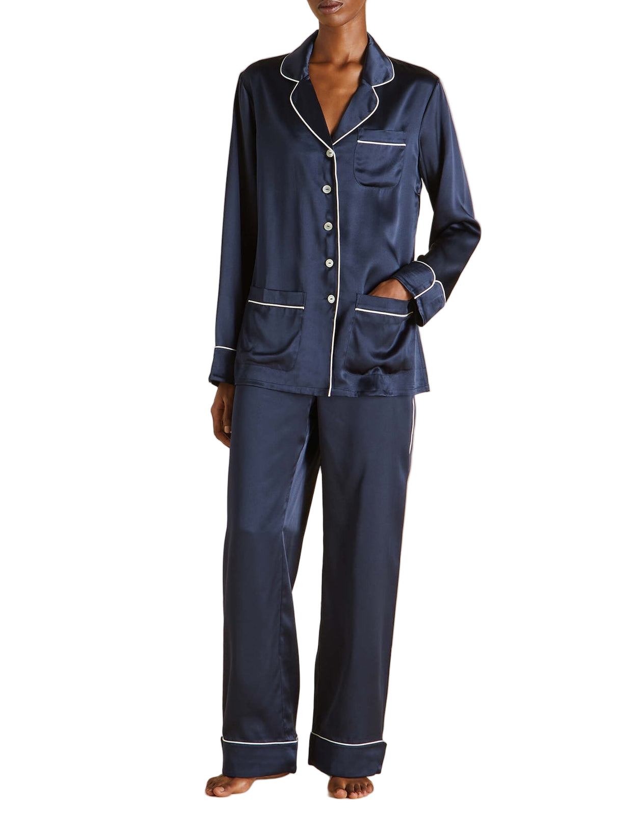 A person models Olivia von Halle’s Coco Silk Pajamas in navy with white piping—featuring a long-sleeve button-up top and matching pants—standing barefoot against a white background.