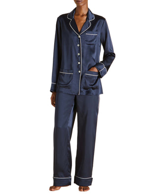 A person models Olivia von Halle’s Coco Silk Pajamas in navy with white piping—featuring a long-sleeve button-up top and matching pants—standing barefoot against a white background.