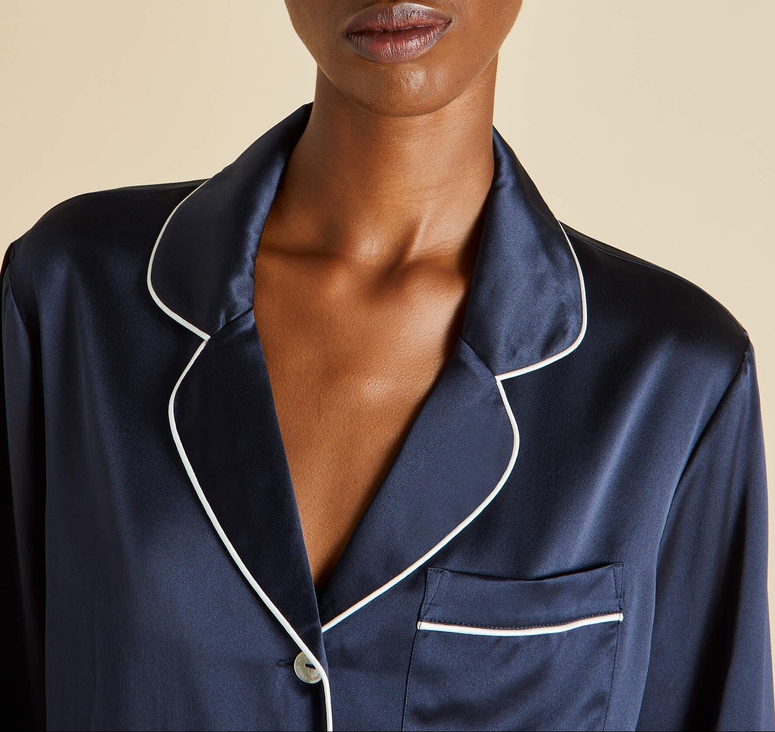 Close-up of a person wearing the Olivia von Halle Coco Silk Pajama top in navy with white piping and a chest pocket, set against a plain beige background.
