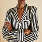 Person wearing an Olivia von Halle Lila Silk Pajamas top with black and white vertical stripes, a collar, and chest pocket, standing with arms crossed to showcase the timeless elegance of this classic pajama style.