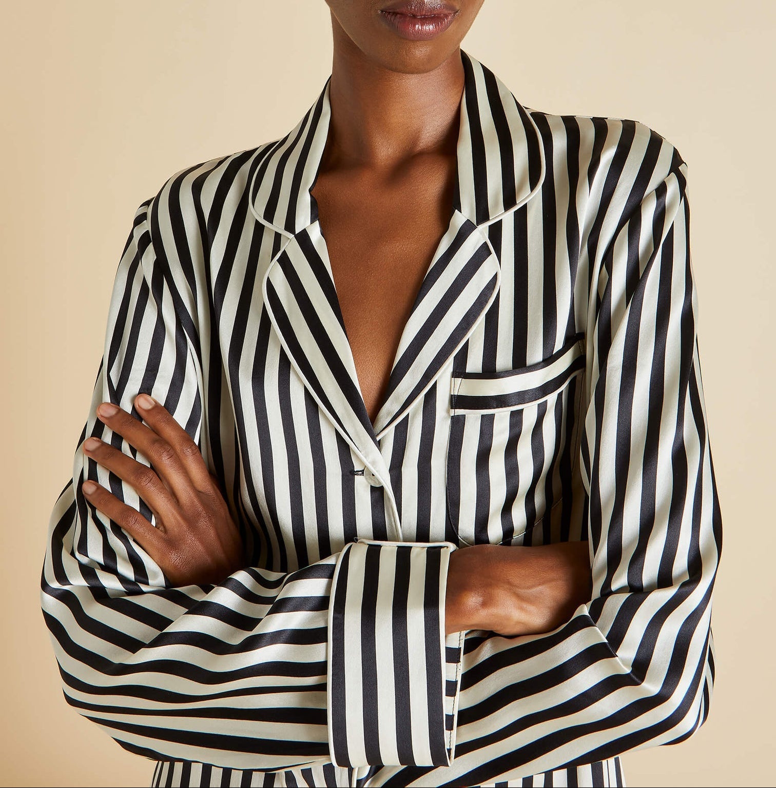 Person wearing an Olivia von Halle Lila Silk Pajamas top with black and white vertical stripes, a collar, and chest pocket, standing with arms crossed to showcase the timeless elegance of this classic pajama style.