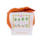 A white box with an orange ribbon and a "Perfectly Palm Beach" label holds the Orange Blossom Candle by Perfectly Palm Beach, inspired by Lilly Pulitzer for a luxurious Palm Beach touch.