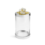 The Orchid Canister by Michael Aram features a clear cylindrical glass design, a silver-toned lid, and a white-and-gold floral accent on top, embodying subtle luxury while reflecting purity and grace.