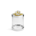 The Michael Aram Orchid Canister features a clear cylindrical jar with a shiny silver lid, topped by a white flower and gold branch from the Orchid Collection—an elegant display of luxury and grace against a plain white background.