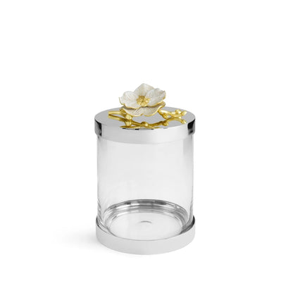 The Michael Aram Orchid Canister features a clear cylindrical jar with a shiny silver lid, topped by a white flower and gold branch from the Orchid Collection—an elegant display of luxury and grace against a plain white background.