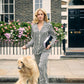Wearing Olivia von Halle’s Lila Silk Pajamas, a woman walks her long-haired dog past a flower-adorned brick townhouse, holding a magazine and folder on a city sidewalk.