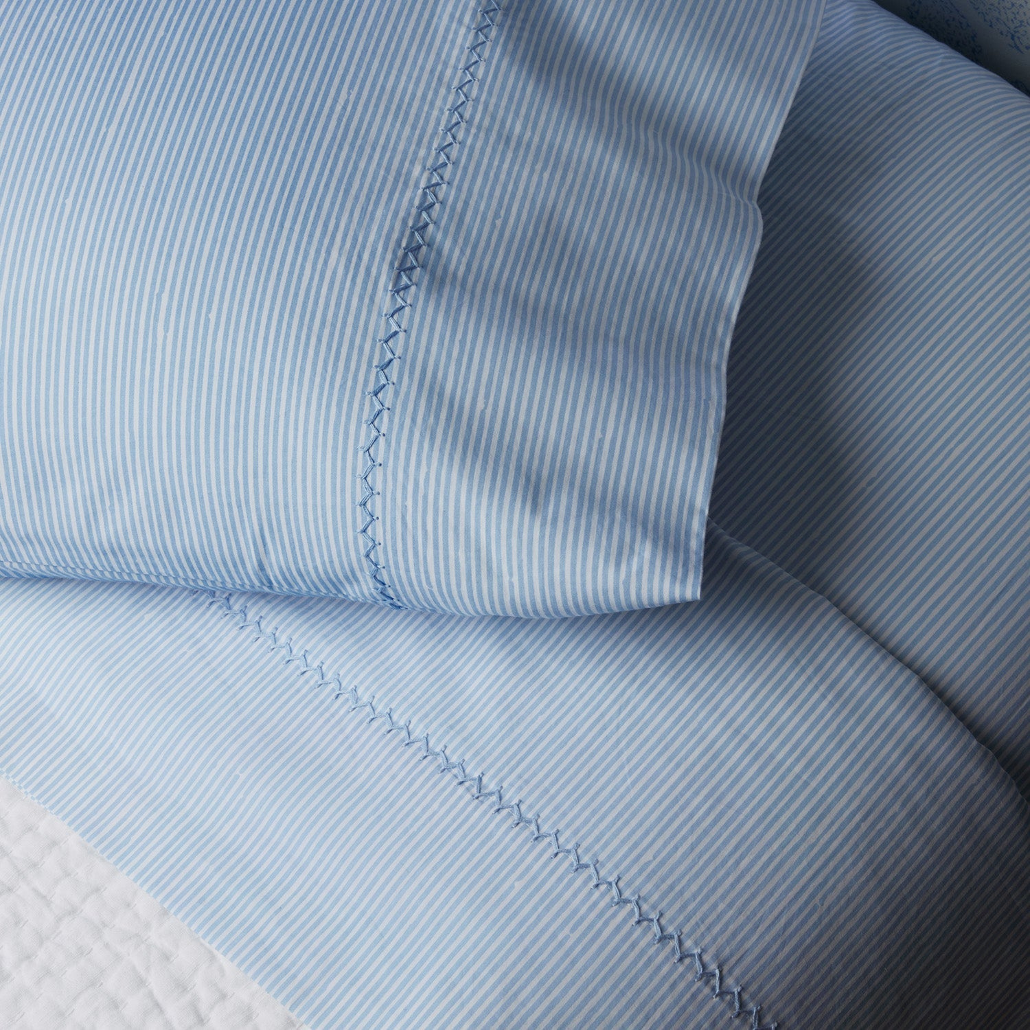 Close-up of a neatly made bed featuring the John Robshaw Nandi Bedding Collection, with light blue and white pinstriped sheets and pillowcases highlighted by decorative stitching, complemented by hand-quilted cotton quilts for an added touch of comfort.