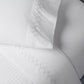 The John Robshaw Stitched Lavender Sheet Set features white organic cotton sheets and pillowcase with light purple zigzag embroidery near the edges, neatly arranged on a bed.