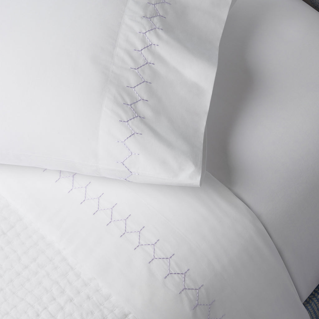 The John Robshaw Stitched Sheet Set in Lavender includes white organic cotton percale bed linens with exquisite purple zigzag hand embroidery on both the pillowcase and top sheet.