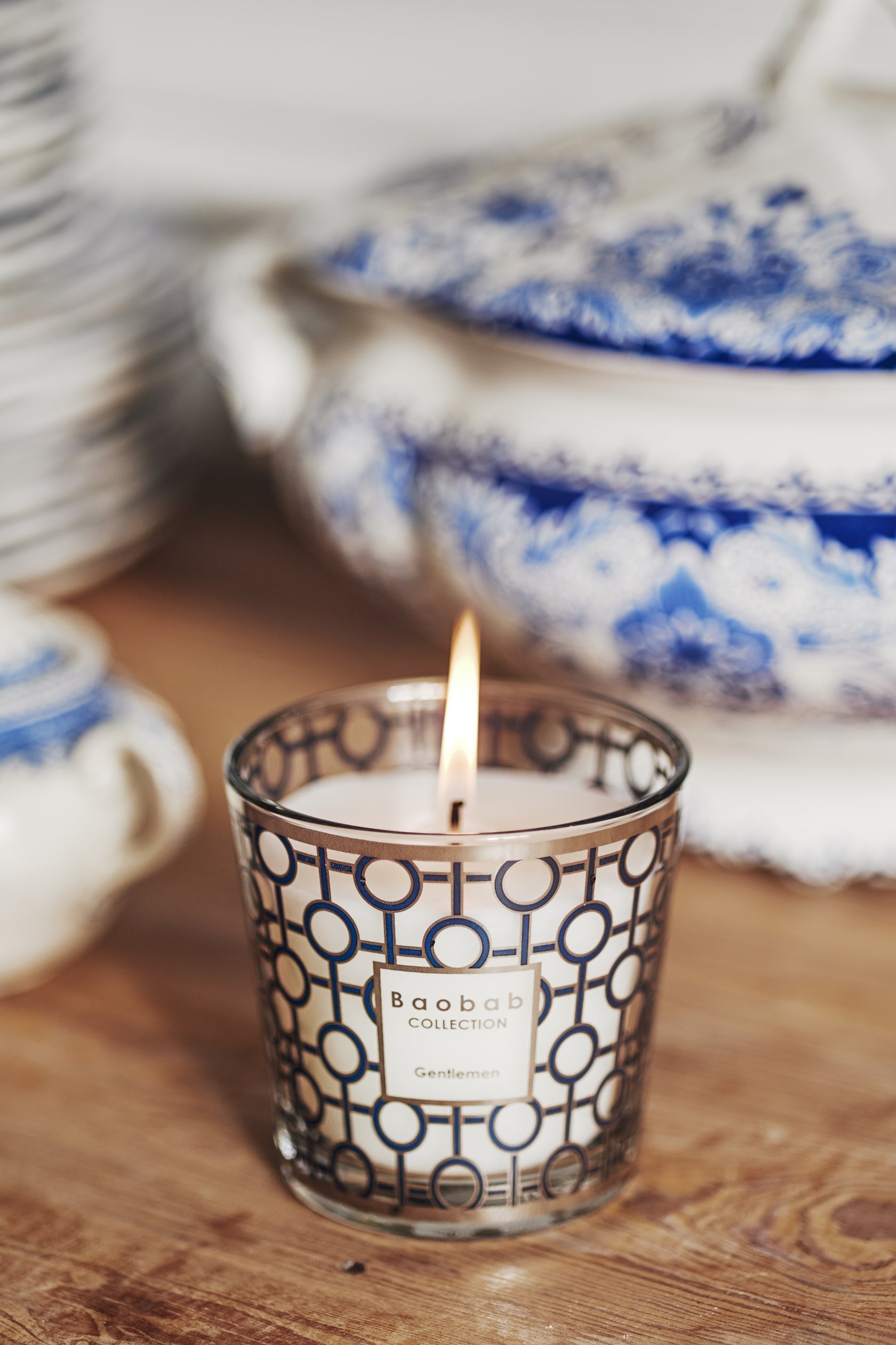 A Baobab Collection Gentlemen Body Wellness candle with a masculine scent rests on a wooden surface beside blue and white ceramic dishes.