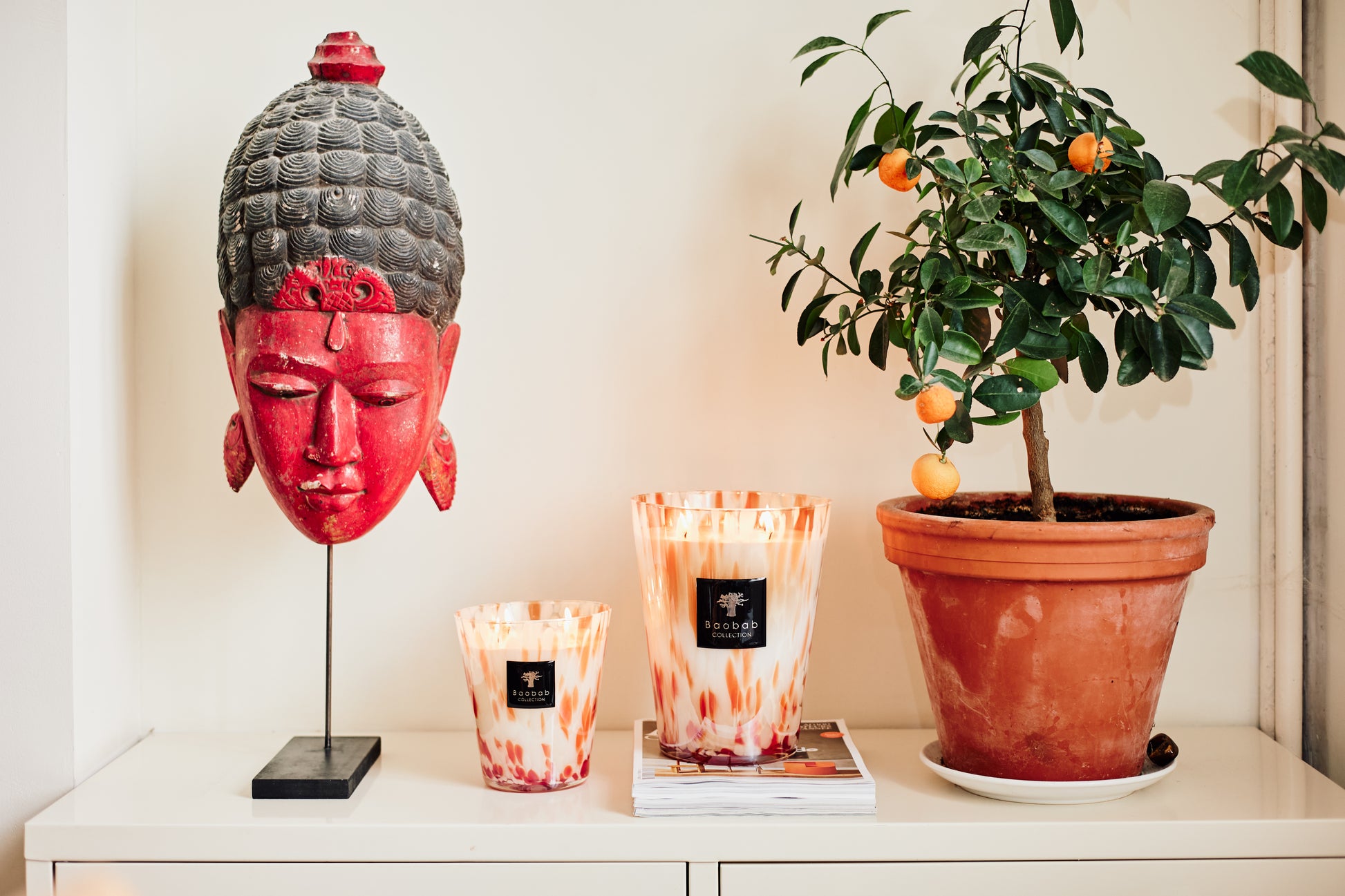 A red decorative Buddha head, a Baobab Collection Pearls Coral Candle, and a potted orange tree are arranged on a white cabinet beside stacked magazines.