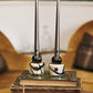 Two Baobab Collection Candle Holder Twins, crafted from exquisite hand-blown glass, rest elegantly on stacked books, while a small bell adds charm to the foreground. The blurred interior in the background enhances the allure of these decorative objects.