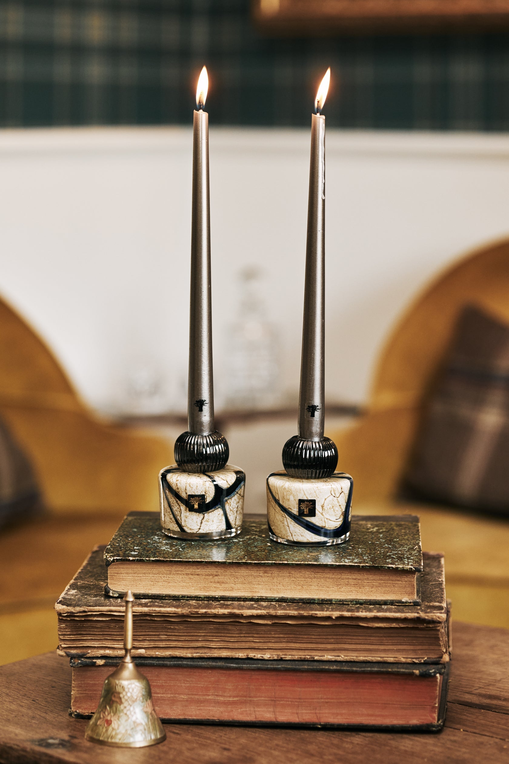 Two Baobab Collection Candle Holder Twins, crafted from exquisite hand-blown glass, rest elegantly on stacked books, while a small bell adds charm to the foreground. The blurred interior in the background enhances the allure of these decorative objects.