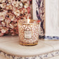 A lit Baobab Women Body Wellness Collection candle by Baobab sits in a decorative glass holder on an ornate white ceramic surface, releasing a floral scent with dried flowers in the background.
