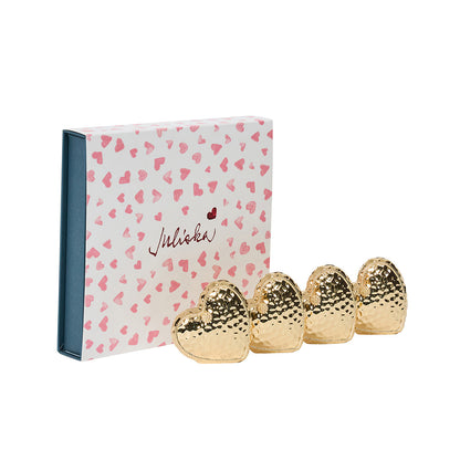 A box with a pink heart pattern and "Miraki" beside four gold, hammered-finish heart-shaped napkin rings from Juliska’s Berry & Thread Heart Napkin Ring and Place Card Holder Collection.