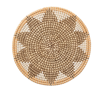 A round, woven seagrass basket tray with a tan, brown, and white starburst pattern pairs perfectly with Kim Seybert’s Majorelle Placemat.