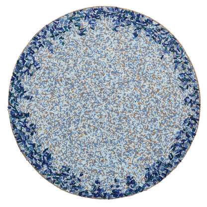 The Kim Seybert Lagoon Placemat showcases a circular mosaic design with a densely patterned blue and brown center accented by cobalt blue beads, bordered by deep ocean-inspired blue and green tones.