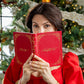 A woman in a red dress holds the Juliska Naughty/Nice Embossed Journal in front of her face, standing by a decorated Christmas tree.